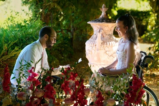 La mariée, assise dans son fauteuil, partage un moment tendre avec le marié dans le jardin.